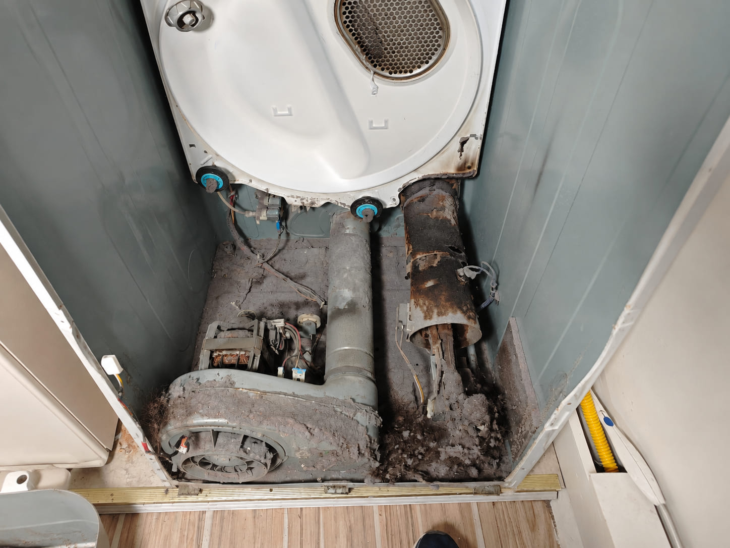 Inside a dryer cabinet with the drum removed — the blower housing and heater duct buried in lint, the duct itself rusted through and scorched