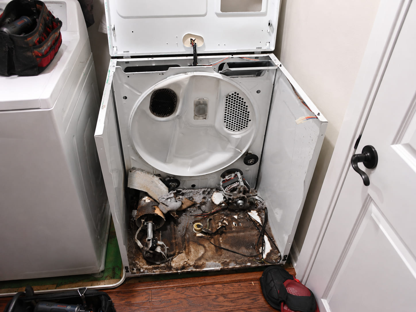 The base of a dryer with the drum lifted out — the slack drive belt draped over the blower housing, the motor pulley and idler wheel beside it