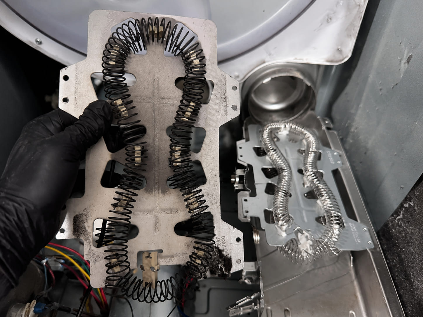 A technician holding a burnt-out dryer heating element, its coil blackened, beside the bright new element already fitted in the housing