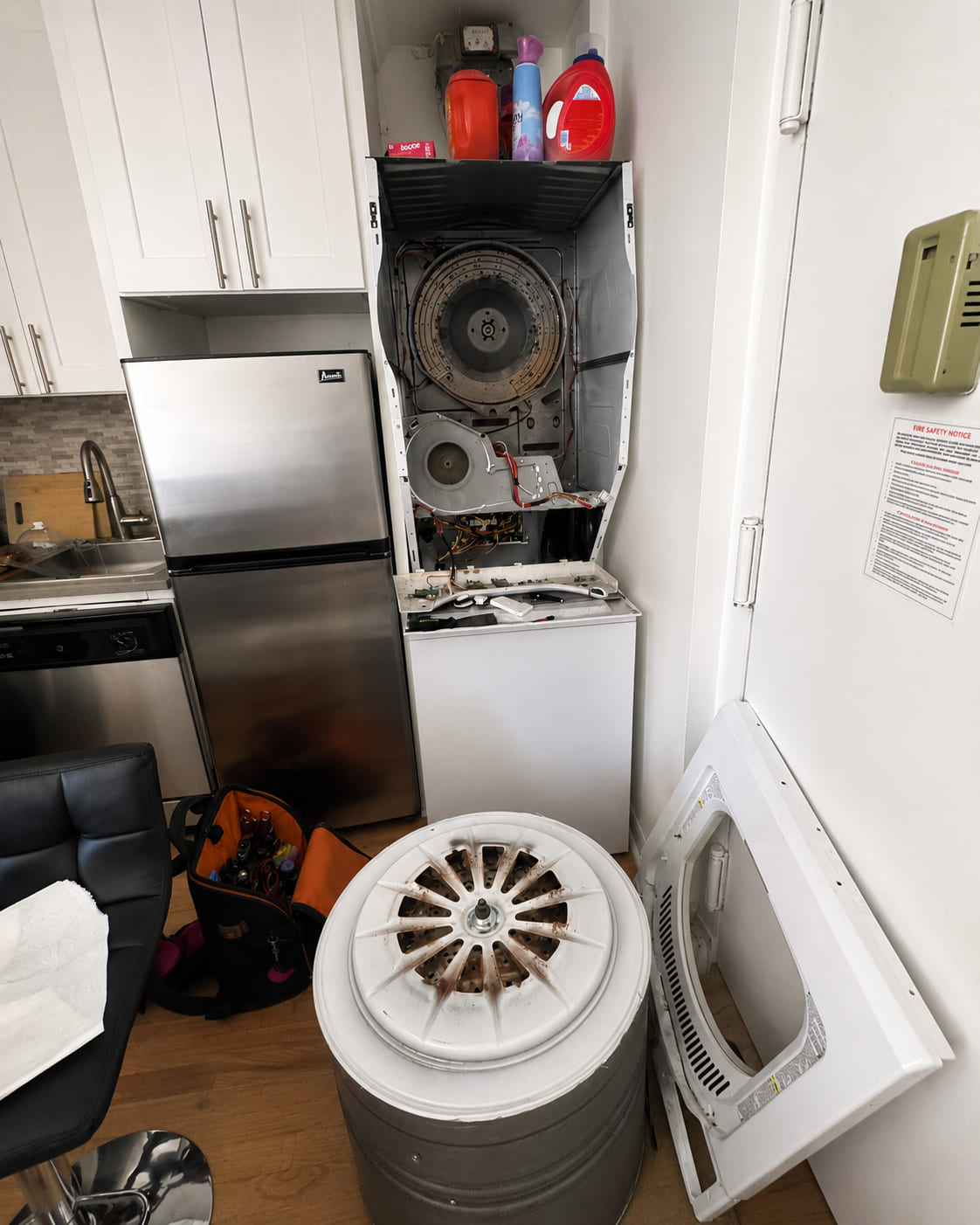 A stacked laundry unit opened up mid-repair, the dryer drum lifted out and standing on the floor beside the removed front panel and a technician's tool bag