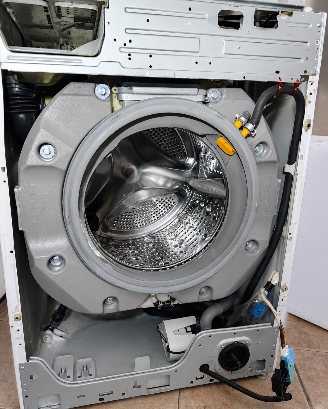 A front-load washing machine with its front panel removed mid-repair, exposing the outer tub, door gasket and drum